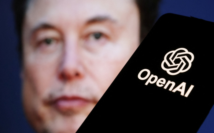 Elon Musk's face blurred in the background while the OpenAI logo appears sharply on a phone screen in the foreground, representing the tense relationship between major players in the AI industry